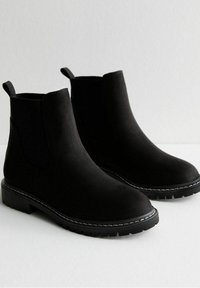 Bottes Chelsea en daim noir avec panneaux latéraux élastiques, bout arrondi et semelle en caoutchouc texturée. Coutures blanches remarquables le long de la semelle.