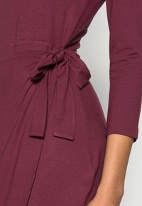 Vestido burgundy com mangas três-quartos, apresentando um cinto de cintura atado. O tecido é suave, com uma textura subtil e um corte descontraído.