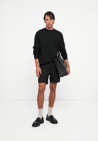 Zwarte lange mouwen shirt, zwarte shorts, beige sokken en zwarte loafers. Het model houdt een zwarte handtas vast. Eenvoudig, minimalistisch ontwerp.