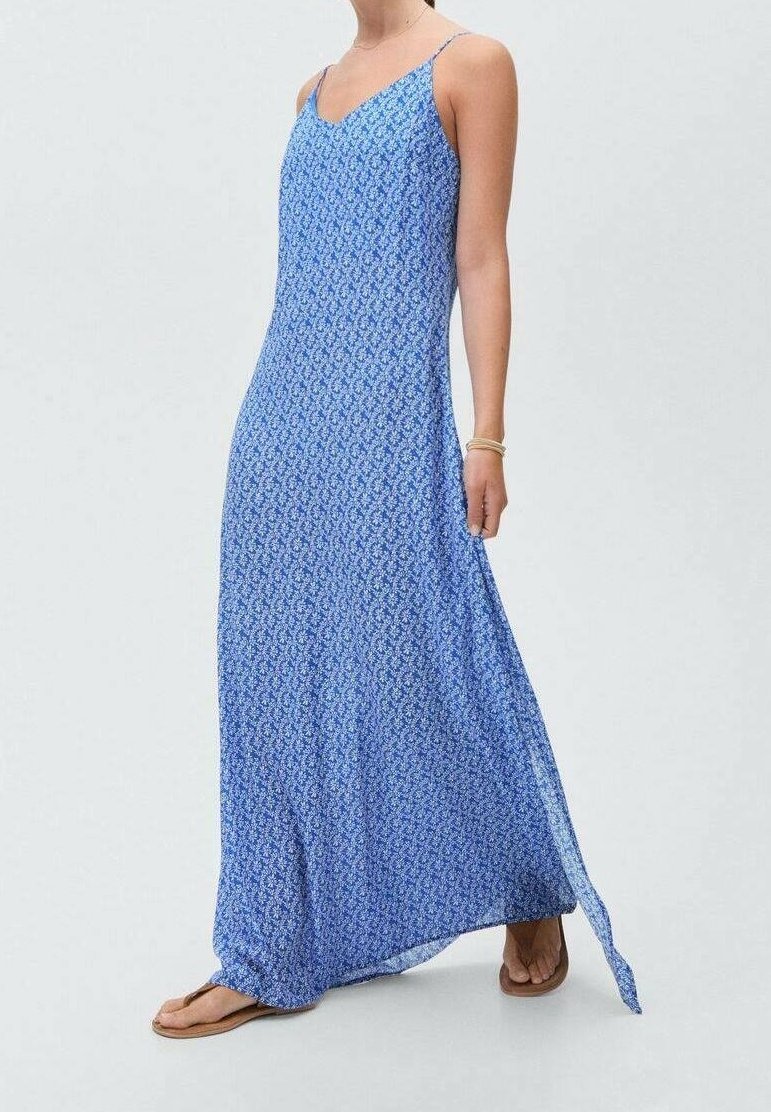 Femme portant une robe maxi bleue sans manches à petits motifs floraux blancs et des sandales marron, debout devant un fond uni.