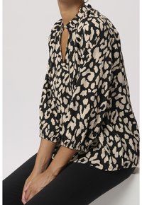 Femme portant un chemisier à motif abstrait noir et beige avec des manches bouffantes 3/4, un col à volants, et un pantalon noir, assise les mains posées sur les genoux.