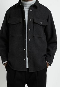 Mörkgrå ullskjortjacka med två bröstfickor, tryckknappsstängning och en texturerad yta. Bärs över en ljusgrå polotröja.