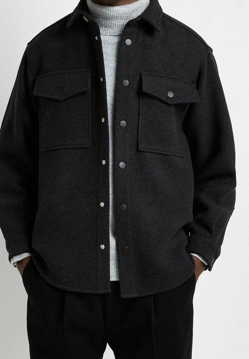 Mörkgrå ullskjortjacka med två bröstfickor, tryckknappsstängning och en texturerad yta. Bärs över en ljusgrå polotröja.