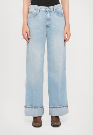Persona che indossa pantaloni a gamba larga di jeans azzurri chiari con grandi risvolti e stivali marroni, in piedi davanti a uno sfondo bianco semplice.