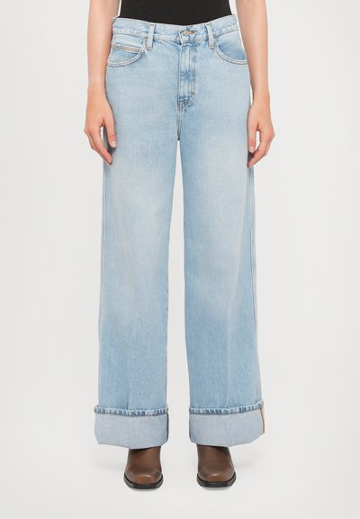 Personne portant un jean large bleu clair avec de grands revers et des bottes marron, se tenant sur un fond blanc uni.