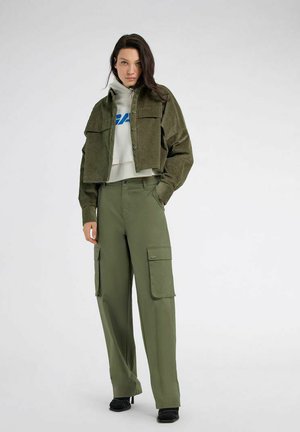 Donna che indossa una giacca corta in velluto a coste verde oliva, felpa bianca con scritta blu, pantaloni cargo verdi oliva e tacchi neri, in piedi su sfondo bianco.