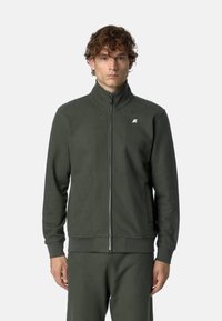 Giacca in fleece verde scuro con colletto alto, zip frontale e tasche laterali. Presenta un piccolo logo bianco sul petto. Tessuto liscio.