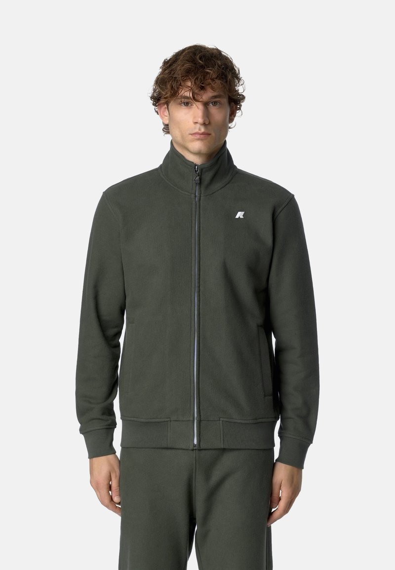 Giacca in fleece verde scuro con colletto alto, zip frontale e tasche laterali. Presenta un piccolo logo bianco sul petto. Tessuto liscio.