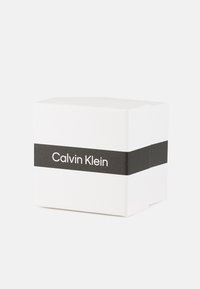 Caja de cartón blanca con una banda horizontal negra etiquetada "Calvin Klein", que presenta un diseño minimalista y una textura suave.