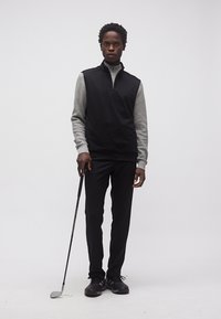 Άνδρας που στέκεται με ένα γκολφ ρόπαλο, φορώντας μαύρο γιλέκο χωρίς μανίκια πάνω από γκρι μακρυμάνικο πουκάμισο, μαύρο παντελόνι και μαύρα παπούτσια σε απλό φόντο.