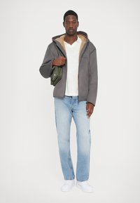 Grå fleecejacka med beige foder, svart dragkedja, vit t-shirt, ljusblå denimjeans och vita sneakers. Grön väska i handen.