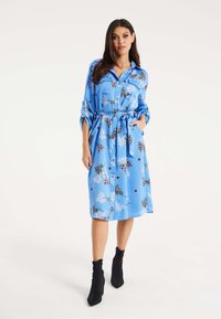 Blue, floral-patterned dress made of satin with a collared neckline, tied waist, and button-down front. Styled with black ankle boots.