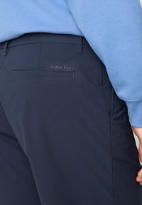 Mørkeblå bukser med en glat tekstur, der har en baglomme og et diskret Calvin Klein-logo. En lys blå topp kan ses over.
