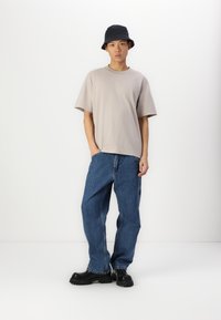 Beige pamut póló, laza szabással; kék denim bő nadrág; fekete vödrös kalap; tömör fekete cipők texturált talppal. Egyszerű, hétköznapi öltözet.