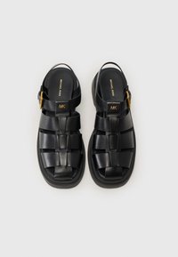 Sorte lærsandaler med flettede stropper, åpen bak og en gullspenne. Flat såle med lett polstring for komfort.