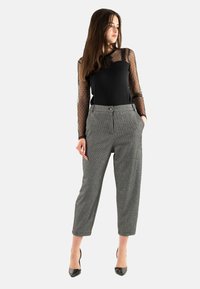 Zwarte top met doorschijnende stippenmouwen, grijs geruite broek met hoge taille en zakken, in combinatie met zwarte puntige hakken.