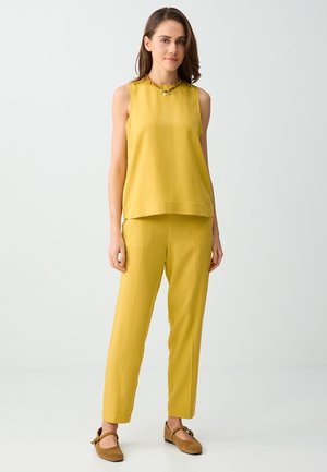 Femme portant un haut jaune sans manches et un pantalon jaune assorti, des chaussures plates marron et un collier de perles sur un fond clair uni.