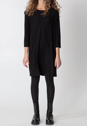 Femme aux cheveux bouclés portant une robe noire jusqu'aux genoux, des leggings noirs et des bottines noires, debout devant un fond blanc uni.