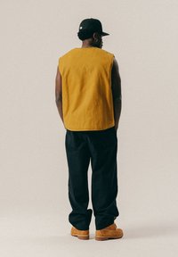 Mies, joka seisoo selin, päällään sinapinvärinen hihaton paita, musta lippalakki, mustat housut ja vaaleat työsaappaat yksinkertaisella taustalla.