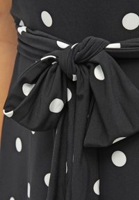 Musta mekko, jossa on valkoisia pilkkuja, ja vyötäröllä on sidottu kaulanauha, joka on valmistettu pehmeästä kankaasta. Tekstuuri on sileä ja kuvio jakautuu tasaisesti.