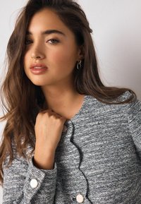 Giacca grigia con trama scura e davanti smerlato, con grandi bottoni rotondi e manica con polsino. Modella che tiene delicatamente il colletto.