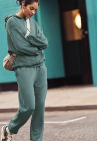 Lysegrøn sweatshirt og matchende joggers lavet af blødt stof. Outfittet har en afslappet pasform. En beige crossover taske bæres.