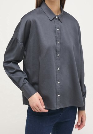 Vrouw draagt een loszittend, donkergrijs overhemd met lange mouwen en knopen, gecombineerd met donkerblauwe spijkerbroek, hand bij heup, neutrale achtergrond.