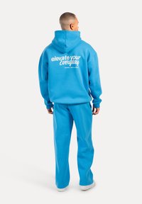 Haut à capuche bleu et pantalon de jogging assorti. Design avec capuche et poche avant. Le dos présente un texte blanc : "élever votre quotidien." Matière douce de sweatshirt.