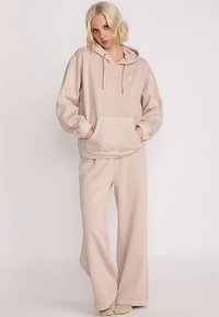 Lichtroze bijpassende hoodie en broekset gemaakt van zachte stof. De hoodie heeft een buidelzak en een trekkoord; de broek heeft wijde pijpen.