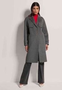 Grau wollener Mantel mit gerader Silhouette, zwei Vordertaschen und Reverskragen. Kombiniert mit dunklen Jeans und cremefarbenen Schuhen.