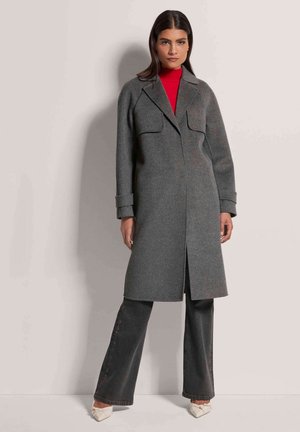 Grijze wollen jas met een rechte silhouet, twee voorzakken en een neklap. Combinatie met donkere spijkerbroek en crèmekleurige schoenen.