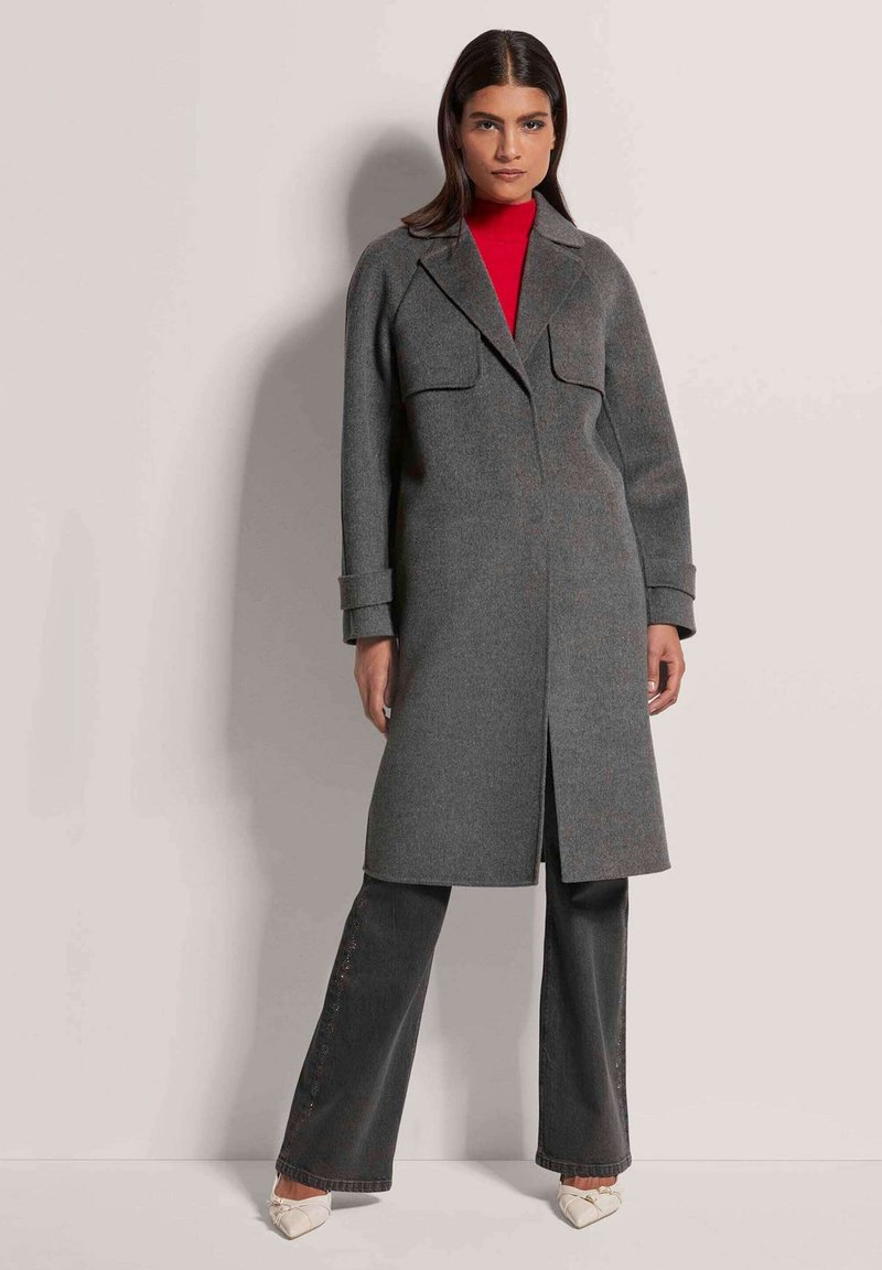 Grau wollener Mantel mit gerader Silhouette, zwei Vordertaschen und Reverskragen. Kombiniert mit dunklen Jeans und cremefarbenen Schuhen.