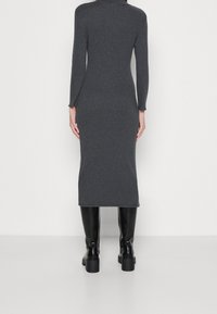 Donna che indossa un vestito midi a maniche lunghe grigio scuro e stivali neri alla coscia con tacco, in piedi di fronte a un muro bianco.