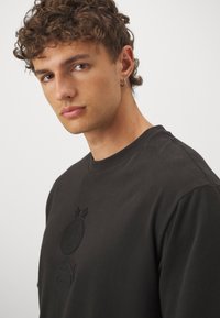 Svart bomullst-shirt med rund halsringning, präglad logotyp med stjärnor i mitten och en subtilt märkt design i botten.