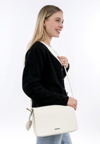 Weißes Leder-Crossbody-Bag mit einer genarbten Textur, silbernem Zubehör und einem herzförmigen Anhänger. Einfaches, rechteckiges Design mit verstellbarem Riemen.