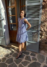 Žena v modro-bielych pruhovaných šatách a čiernych podpätkoch stojí na slnku pri drevených dverách so svetlomodrými okenicami na kamennej podlahe.