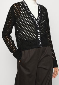 Svart cardigan med öppen stickning, v-ringning, långa ärmar och ett logoband längs framsidan, kompletterad med ribbad kant och muddar.