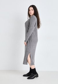 Rochie tricotată gri cu mâneci lungi, fante laterale și textură cu nervuri. Asortată cu pantofi negri și șosete. Siluetă simplă, dreaptă.