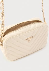 Krémová taška prošívaná vzorem s kovovým zlatým řetízkovým popruhem a detailem loga. Má zapínání na zip a texturovaný povrch s diagonálním prošíváním.