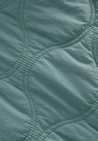 Quiltet sengetæppe i lys teal med bølgede syede mønstre. Stoffet har en blød, let krøllet tekstur.