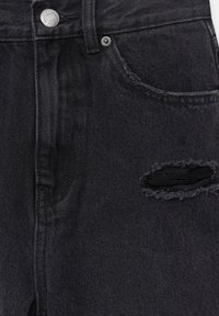 Gros plan sur un jean en denim noir avec un trou déchiré près de la poche avant et des détails visibles de bouton et rivet en métal.