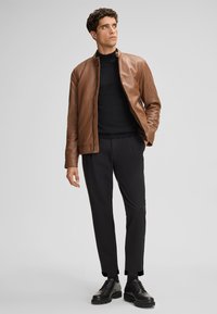 Bruine leren jas met een ronde kraag, gecombineerd met een zwarte trui en op maat gemaakte zwarte broeken. Zwarte veterschoenen maken de outfit compleet.