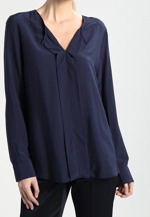 Vrouw met een marineblauwe blouse met lange mouwen, een V-hals en geplooide details, gecombineerd met zwarte broek met textuur.