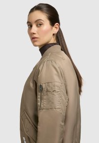Beige Bomberjacke mit einem Reißverschlusstasche am linken Ärmel, geripptem Kragen und einer glatten, glänzenden Textur.