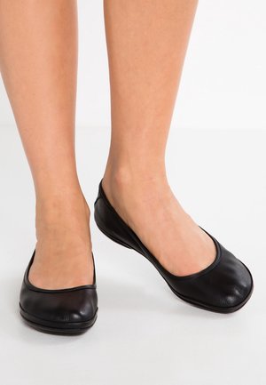 Weibliche Füße, die schlichte schwarze Leder-Ballerinas auf einer weißen Oberfläche tragen.