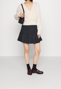 Vaaleanbeige neulepaita, jossa syvä V-pääntie ja resorikuvio. Musta liehuhame, jossa laskokset. Malli käyttää tummia sukkia ja ruskeita loafer-kenkiä.