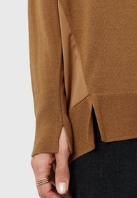 Maglione marrone lavorato a maglia con fessure laterali e polsini a costine. Tessuto morbido, vestibilità rilassata. Ulteriore strato di tessuto leggero visibile sotto.