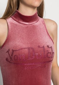 Fajanslös topp i rosa sammet med hög krage. Har "Von Dutch"-logotypen dekorerad med rosa rhinestones ovanför "ORIGINALS." Slät textur.