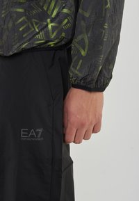 Giacca sportiva nera con parte superiore traslucida e modelli geometrici verdi, abbinata a pantaloni neri con il logo EA7 in grigio.