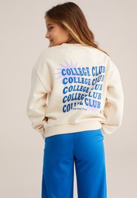 Kremkrāsas džemperis ar daudzkrāsainu „KOLEDŽU KLUBS” uzrakstu un saules grafiku uz aizmugures. Mīksta auduma un ribota aproce. Apvienots ar zilām biksēm.